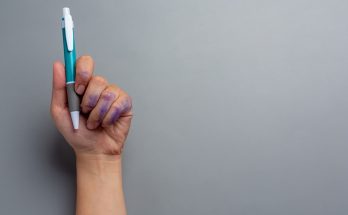Eine Hand hält einen blauen Kugelschreiber, während die Finger von lila Tinte verschmiert sind. Der Hintergrund ist grau und neutral, was den Fokus auf den Stift und die Hand lenkt.