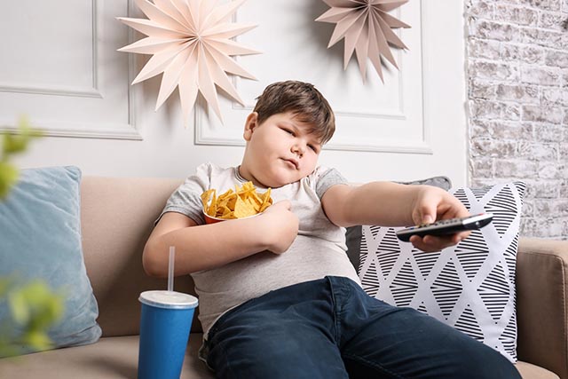 Übergewichtiges Kind sitzt mit Snacks vor dem Fernseher