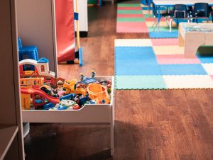 Bunte Spielzeugkiste mit verschiedenen Spielsachen in einem hellen, kinderfreundlichen Raum mit farbigen Teppichfliesen.
