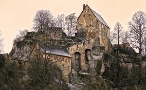 Historische Burganlage mit steinernen Gebäuden, umgeben von Bäumen und Felsen, in sanftem Licht.