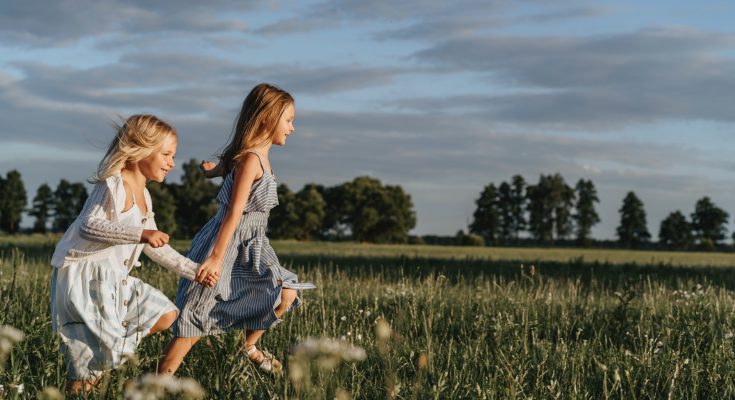 Zwei Mädchen laufen fröhlich über ein blühendes Feld unter einem klaren Himmel. Die Szene strahlt Unbeschwertheit und Freude aus, ideal für Themen wie Kindheit und Naturerlebnisse.