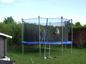 Zwei Kinder springen auf einem runden Trampolin mit Sicherheitsnetz in einem grünen Garten. Im Hintergrund sind Büsche und ein blauer Himmel sichtbar.