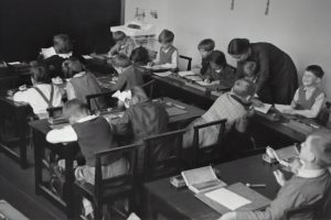 Schwarz-weißes Bild von einer Schulklasse in den 1950er Jahren, in der Schüler an Schreibtischen arbeiten, während ein Lehrer ihnen Anweisungen gibt.