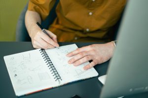 Eine Person schreibt in ein Notizbuch, während sie an einem Schreibtisch sitzt. Auf dem Tisch steht ein Laptop. Die Hand ist deutlich sichtbar, während sie Skizzen oder Notizen anfertigt.