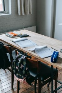 Ein Holztisch in einem modernen Raum, bedeckt mit Notizen, Büchern und Schreibutensilien. Im Hintergrund hängen Stühle mit Rucksäcken. Die Szene vermittelt eine kreative Arbeitsatmosphäre.