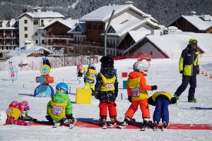 Skifahrende Kinder in bunten Skianzügen auf einem Übungshang, umgeben von schneebedeckten Bergen und modernen Chalets.