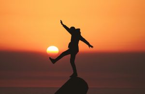 Mann balanciert auf einem Felsen mit ausgestrecktem Bein vor untergehender Sonne, es sieht aus, als ob die Sonne, wie einen Ball, mit dem Fuß trifft.