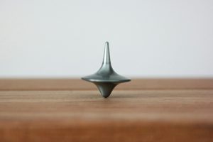 Ein metallener Kreisel steht auf einer Holzoberfläche und dreht sich. Der Kreisel hat eine schlanke, konische Form und glänzt silbern. Ideal für Spiele oder als dekoratives Element.