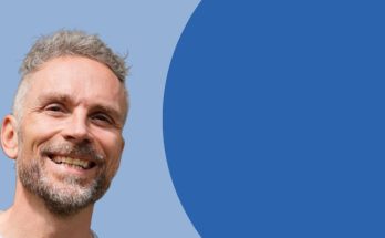 Lächelnder Mann mit grauen Haaren vor einem blauen Hintergrund, der eine freundliche und einladende Ausstrahlung hat. Ideal für Themen wie persönliche Entwicklung oder Coaching.