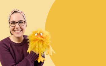 Eine lächelnde Frau mit Brille und einem bunten, gelben Handpuppenmonster auf einem gelben Hintergrund. Sie trägt einen lila Pullover und ein Kopftuch, was eine freundliche und einladende Atmosphäre schafft. Ideal für Inhalte über Kinderunterhaltung oder Puppenspiel.