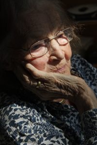 Nahaufnahme einer älteren Frau mit Brille, die nachdenklich wirkt. Ihr Gesicht ist von sanftem Licht beleuchtet, das die Falten und Textur ihrer Haut betont. Sie trägt ein gemustertes Oberteil und hält ihre Hand an das Kinn.