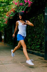 Eine sportliche Frau in einem weißen Tanktop und blauen Shorts läuft auf einem Gehweg, umgeben von grünen Pflanzen und bunten Blumen. Die Szene vermittelt einen aktiven und frischen Lebensstil.