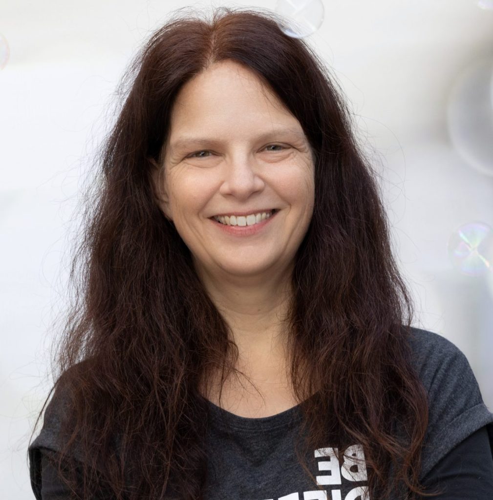 Porträt einer lächelnden Frau mit langen, lockigen Haaren, die ein schwarzes Shirt mit der Aufschrift "BE DIFFERENT" trägt. Der Hintergrund ist unscharf mit blassen Lichtreflexen, was eine freundliche und einladende Atmosphäre schafft.