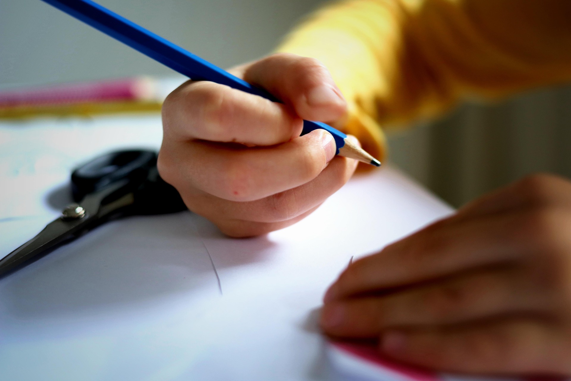 Eine Hand hält einen blauen Bleistift und zeichnet auf ein Blatt Papier, während eine Schere im Hintergrund zu sehen ist.