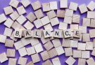 Holzbuchstaben auf lila Hintergrund bilden das Wort "BALANCE". Die Buchstaben sind in einem kreativen Arrangement zwischen leeren Holzplättchen angeordnet, was das Thema Gleichgewicht und Harmonie symbolisiert.