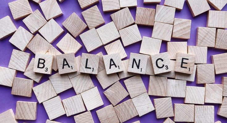 Holzbuchstaben auf lila Hintergrund bilden das Wort "BALANCE". Die Buchstaben sind in einem kreativen Arrangement zwischen leeren Holzplättchen angeordnet, was das Thema Gleichgewicht und Harmonie symbolisiert.