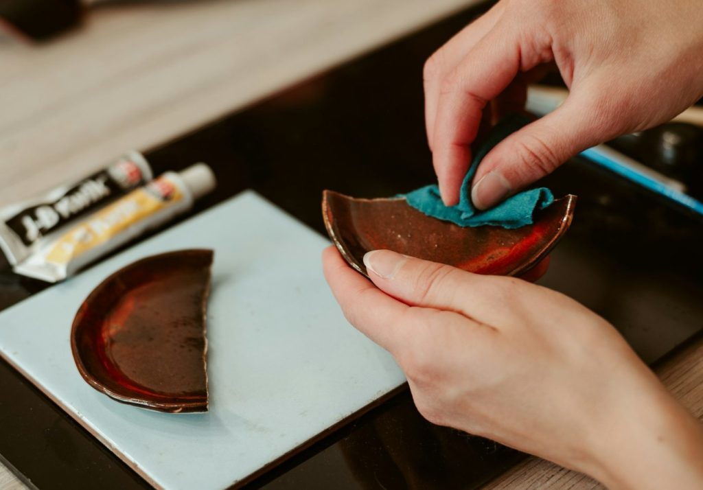 Zwei Hände arbeiten an einem zerbrochenen Schälchen und eine Hand putzt mit einem blauen Lappen einen Teil der zerbrochenen Schale.