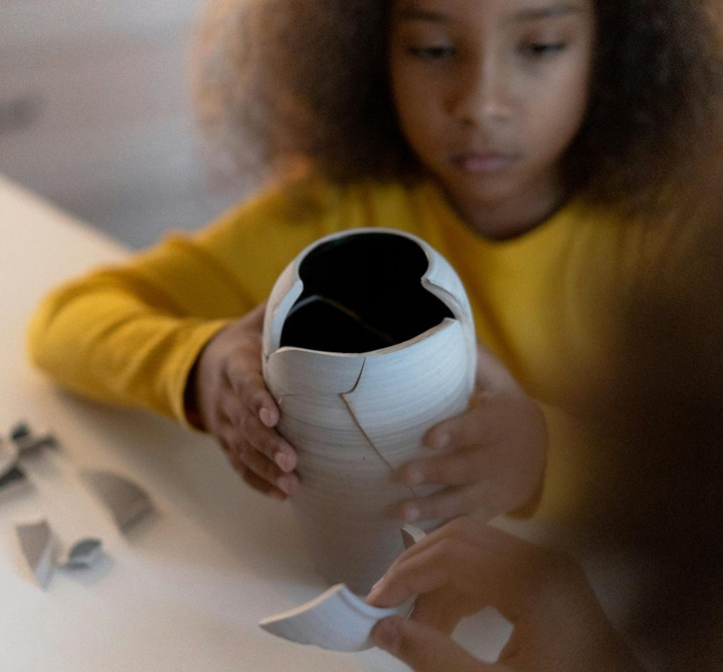 Ein Kind mit lockigem Haar hält eine beschädigte Vase in den Händen und versucht, die zerbrochenen Teile zusammenzufügen. Auf dem Tisch liegen weitere Scherben, die zur Reparatur der Vase verwendet werden sollen.