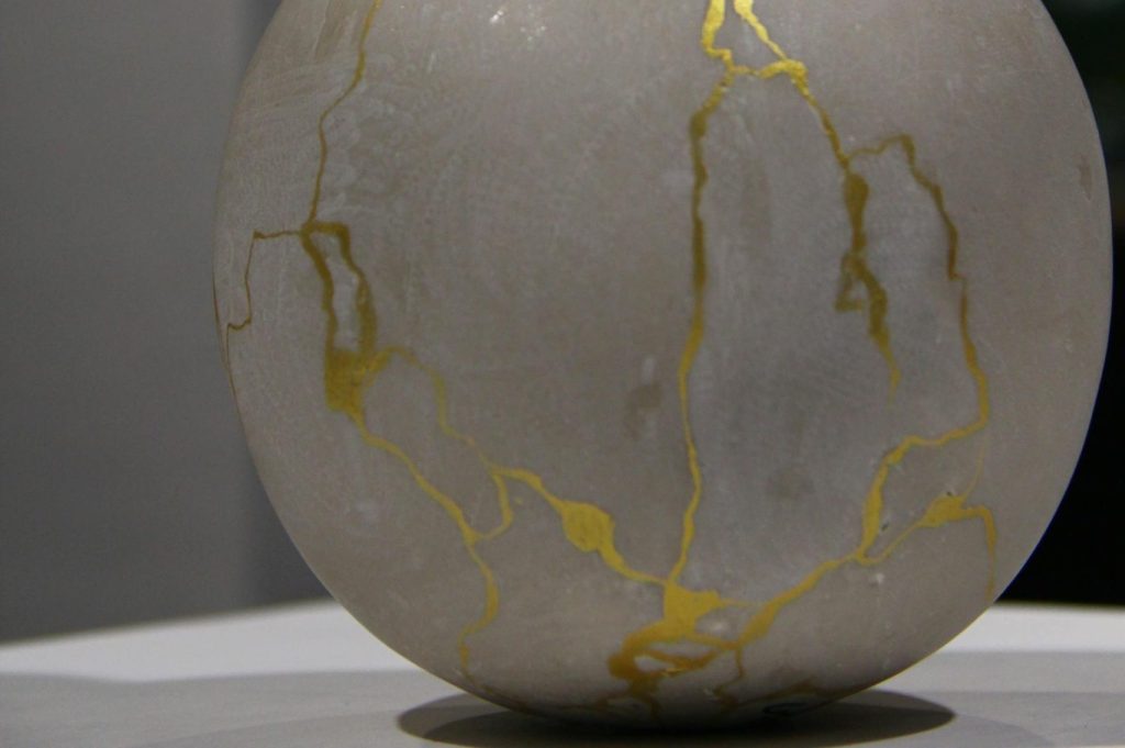 Künstlerische Keramikvase mit goldenen Adern auf einer hellen Oberfläche, präsentiert in einer modernen Ausstellung. Die Vase hat eine elegante, geschwungene Form und hebt sich durch den Kontrast zwischen dem matten Finish und den glänzenden goldenen Details hervor.