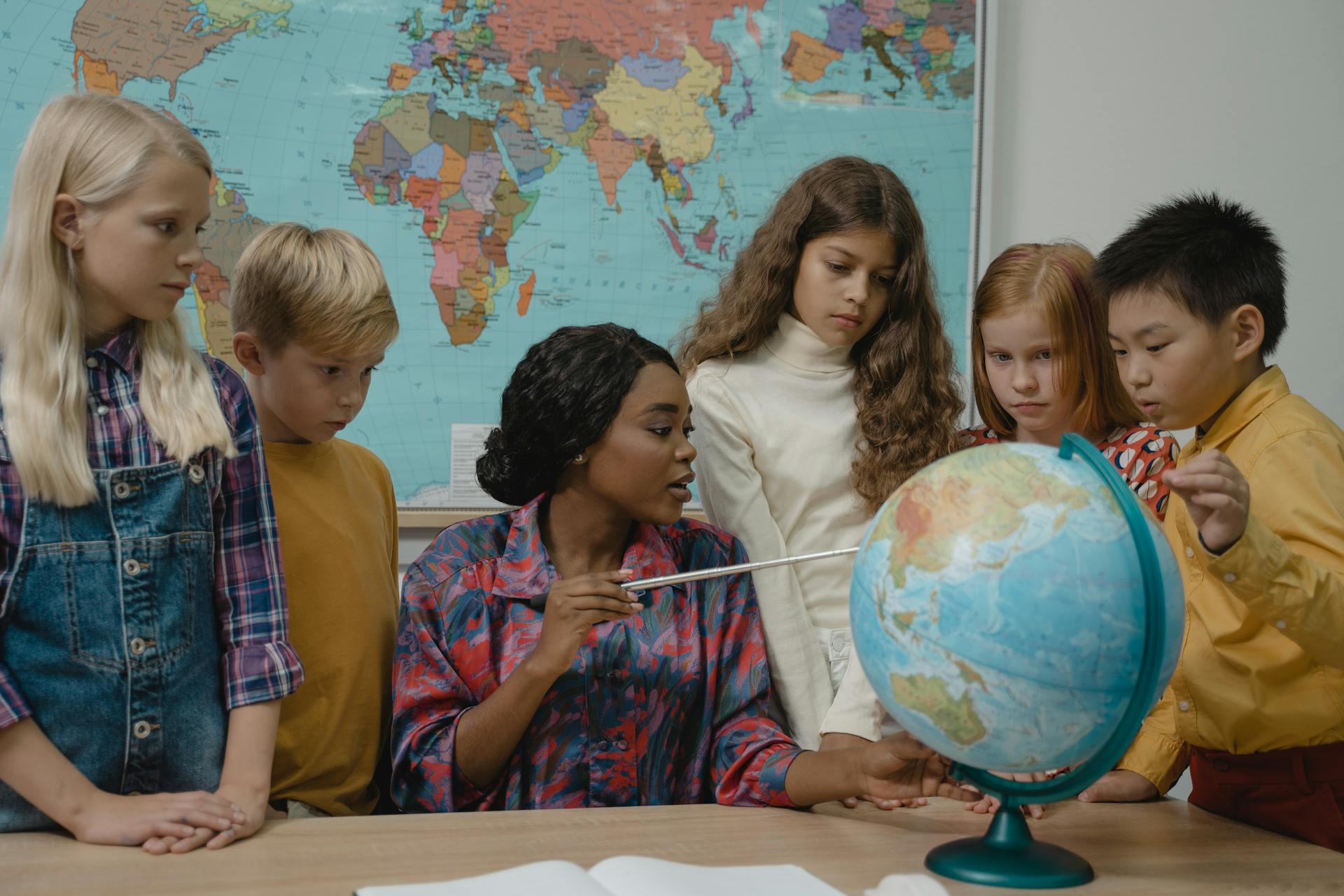 Lehrerin erklärt den Schülern an einem Globus geografische Konzepte, während eine Weltkarte im Hintergrund zu sehen ist. Sechs Kinder unterschiedlichen Alters und Geschlechts zeigen Interesse und Konzentration.