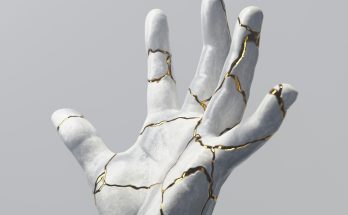Eine stilisierte Hand aus weißem Material mit Rissen, die mit goldenen Linien akzentuiert sind, vor einem grauen Hintergrund. Die Darstellung symbolisiert Zerbrechlichkeit und Schönheit.