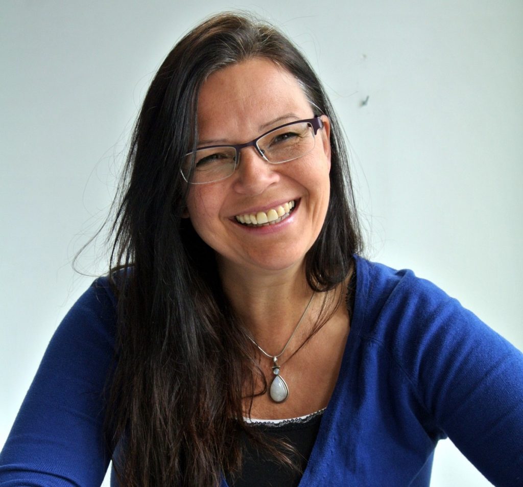 Lächelnde Frau mit langen, dunklen Haaren und Brille, die in einem blauen Pullover vor einem hellen Hintergrund sitzt.