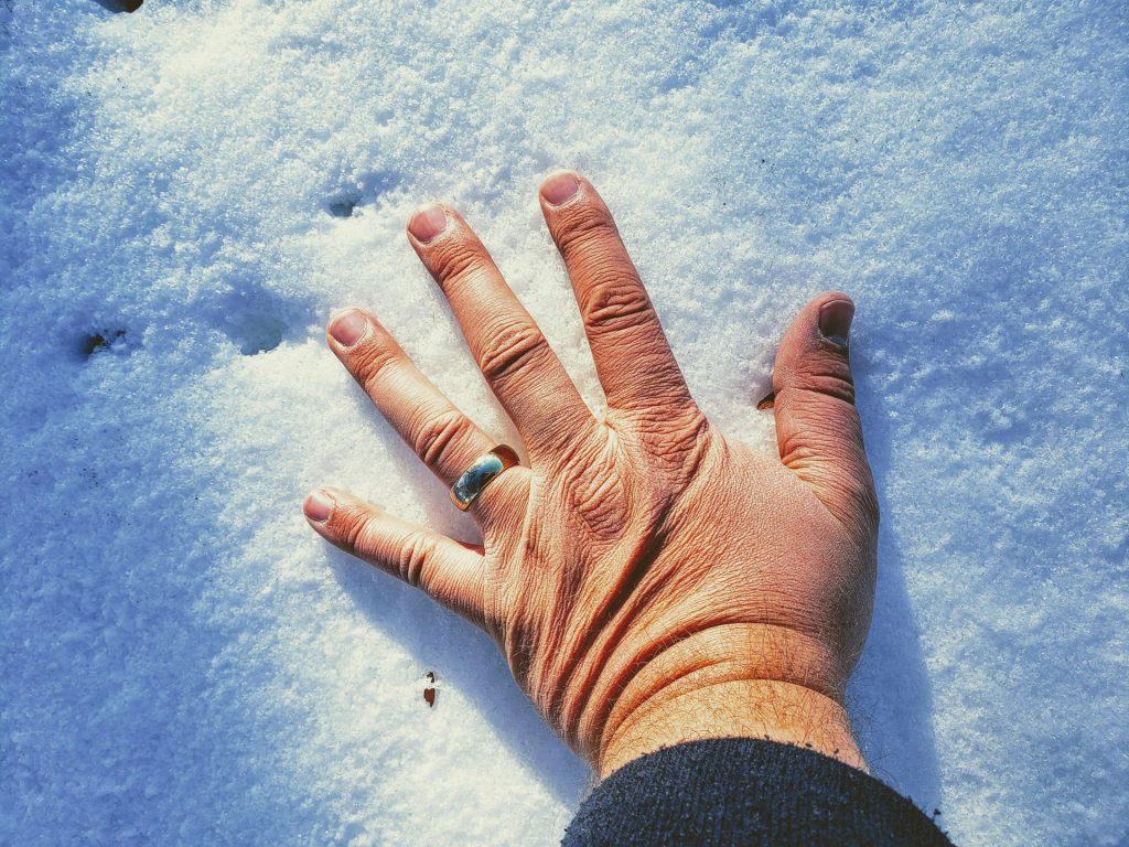 Eine Hand mit einem Ring liegt auf frischem, unberührtem Schnee. Die Textur des Schnees ist deutlich sichtbar, während die Sonne sanft darauf scheint.