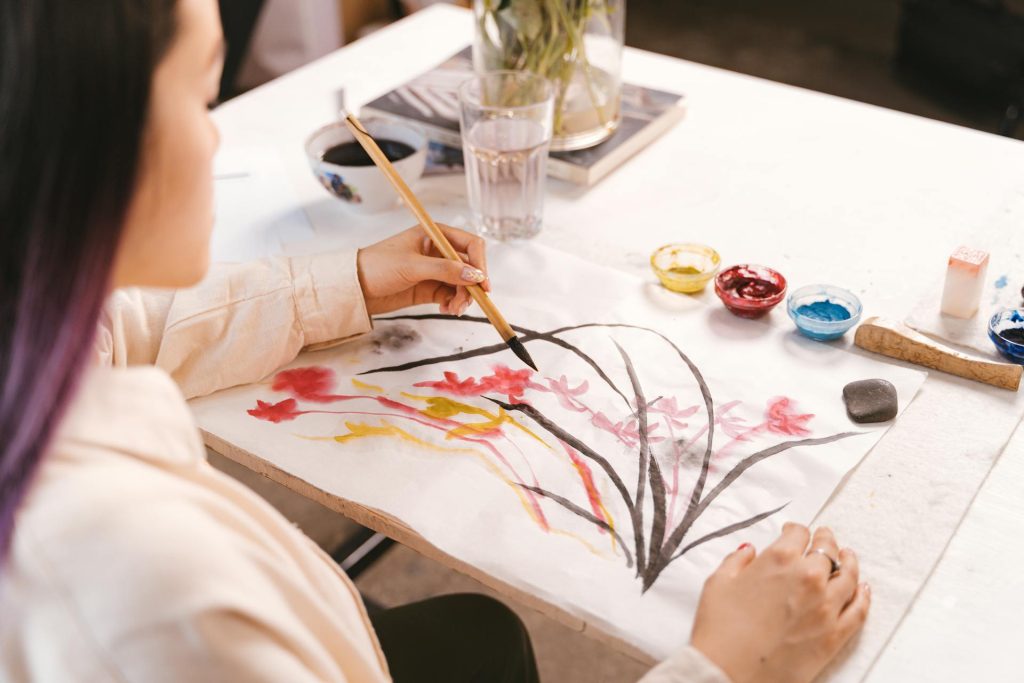 Eine Künstlerin malt mit einem Pinsel auf Papier, während sie bunte Aquarellfarben und Materialien auf dem Tisch um sich hat. Die Darstellung zeigt florale Motive in sanften Farben.