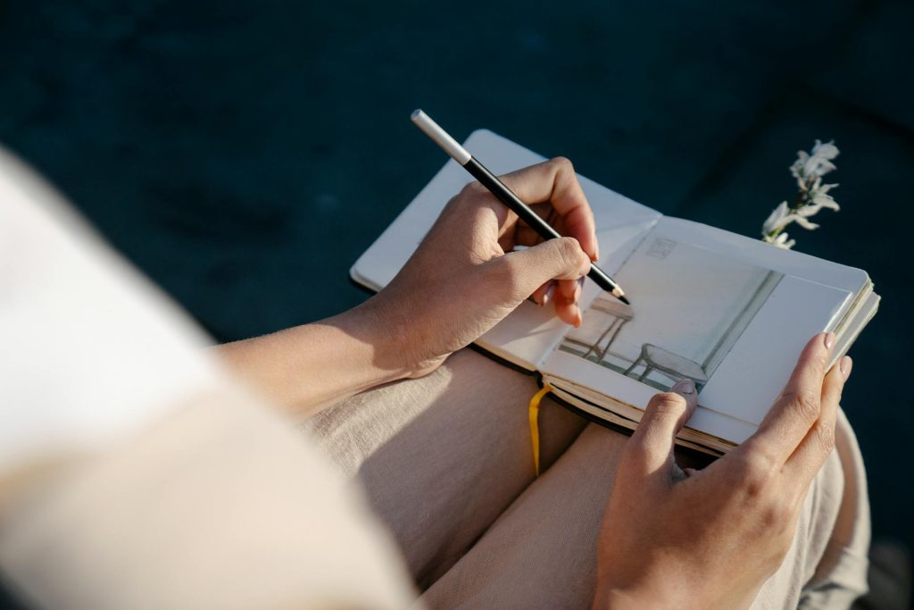 Eine Person zeichnet in einem Skizzenbuch mit einem Bleistift. Die Skizze zeigt einen Stuhl in einem hellen Raum. Der Fokus liegt auf den Händen der Person und dem offenen Buch, während im Hintergrund eine blühende Pflanze zu sehen ist.