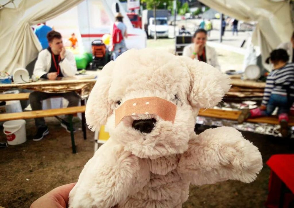Plüschhund mit einem Pflaster auf der Schnauze, gehalten von einer Person. Im Hintergrund sind Menschen und eine Zeltatmosphäre zu sehen, die auf eine Veranstaltung hindeutet.