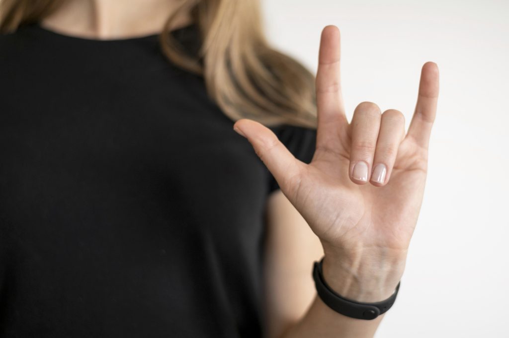 Eine Person zeigt mit der Hand das "I Love You"-Zeichen in Gebärdensprache. Die Hand ist gut sichtbar und die Fingernägel sind gepflegt. Der Hintergrund ist neutral und lenkt nicht ab.