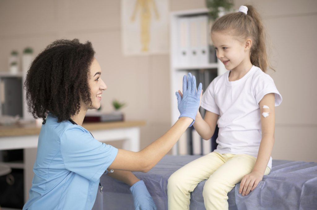 Eine Krankenschwester interagiert freundlich mit einem kleinen Mädchen in einer medizinischen Umgebung. Das Mädchen sitzt auf einer Liege und hat ein Pflaster am Arm, während die Krankenschwester mit Handschuhen die Hand des Mädchens hochhält.