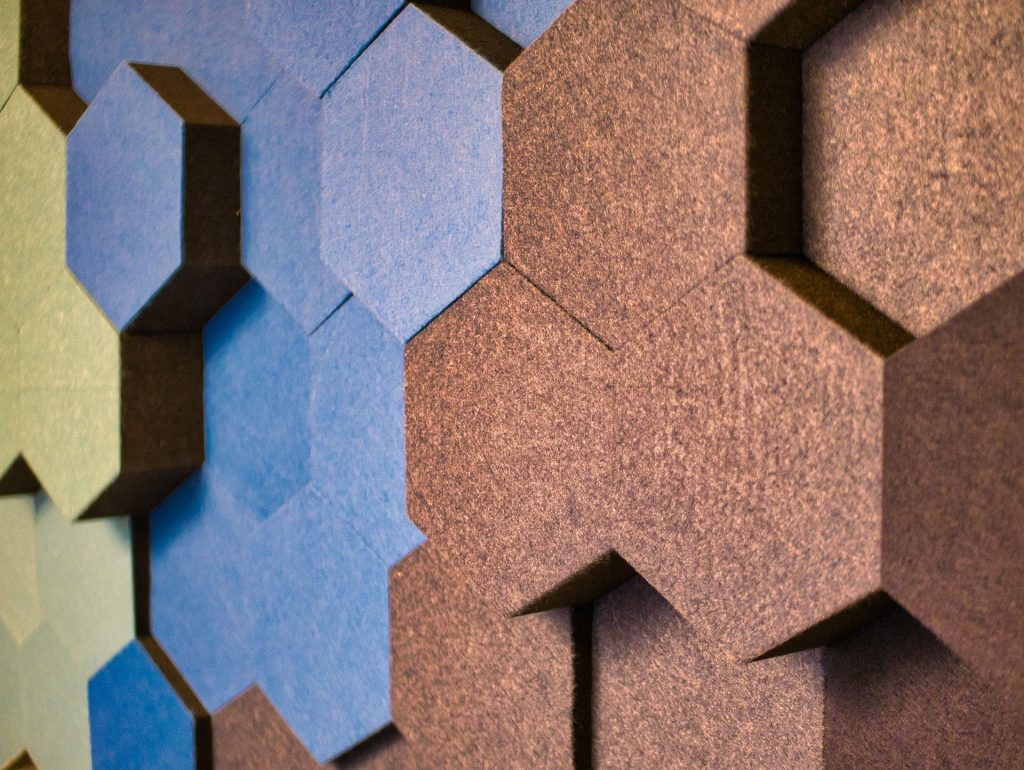 Farbenfrohe Wandverkleidung aus hexagonalen Paneelen in verschiedenen Blau- und Brauntönen. Die Struktur sorgt für eine moderne und ästhetische Raumgestaltung. Ideal für Büros oder kreative Arbeitsumgebungen.