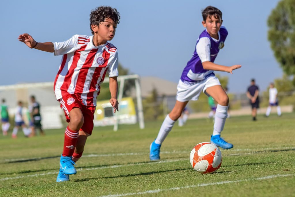 Zwei junge Fußballspieler im Wettkampf auf dem Spielfeld, einer in rot-weißem Trikot und der andere in lila-weißem Trikot, während sie einem Fußball nachjagen.