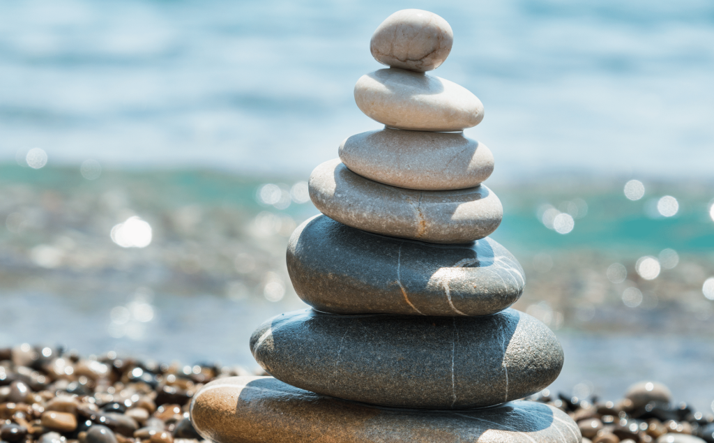 Steinmosaik aus verschiedenen glattpolierten Steinen, sorgfältig aufeinander gestapelt, vor einem ruhigen Wasserhintergrund. Die Komposition vermittelt ein Gefühl von Ruhe und Balance, ideal für Entspannung und Meditation.