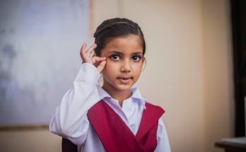 Ein kleines Mädchen mit dunklen Haaren trägt ein weißes Hemd und eine rote Weste. Sie schaut direkt in die Kamera und zeigt mit einer Hand ein Zeichen. Der Hintergrund ist unscharf und zeigt eine Schulumgebung.
