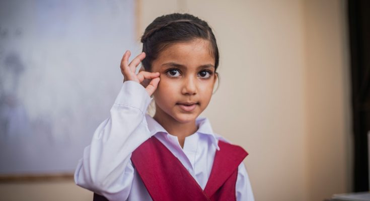 Ein kleines Mädchen mit dunklen Haaren trägt ein weißes Hemd und eine rote Weste. Sie schaut direkt in die Kamera und zeigt mit einer Hand ein Zeichen. Der Hintergrund ist unscharf und zeigt eine Schulumgebung.
