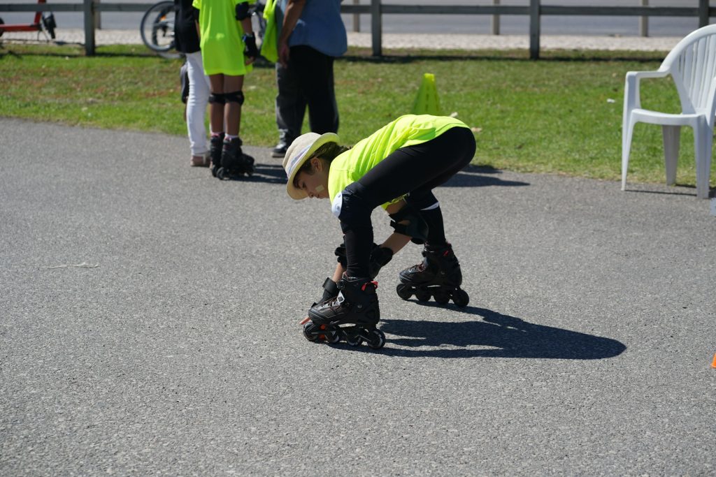 Ein Kind in leuchtend gelber Sportbekleidung und einem Hut kniet auf dem Asphalt, um seine Rollschuhe anzuziehen. Im Hintergrund sind weitere Kinder und Erwachsene zu sehen, die an einer Outdoor-Aktivität teilnehmen. Die Szene spielt sich an einem sonnigen Tag ab, ideal für sportliche Aktivitäten im Freien.