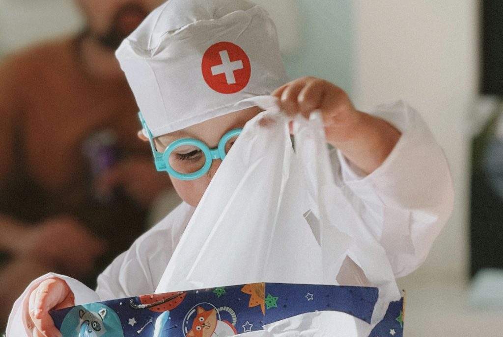 Ein kleines Kind in einem Arztkostüm mit einer roten Kreuzmütze und blauen Brillen öffnet ein Geschenk, umgeben von buntem Geschenkpapier mit Tiermotiven im Weltraum. Im Hintergrund ist ein Erwachsener zu sehen.