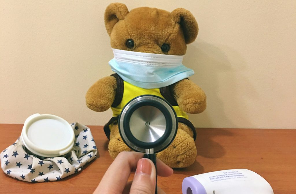 Stoffbär mit Mundschutz sitzt auf einem Tisch, während eine Hand ein Stethoskop hält. Neben dem Bären liegen ein Thermometer und ein Tuch mit Sternen. Das Bild vermittelt eine kinderfreundliche medizinische Atmosphäre.