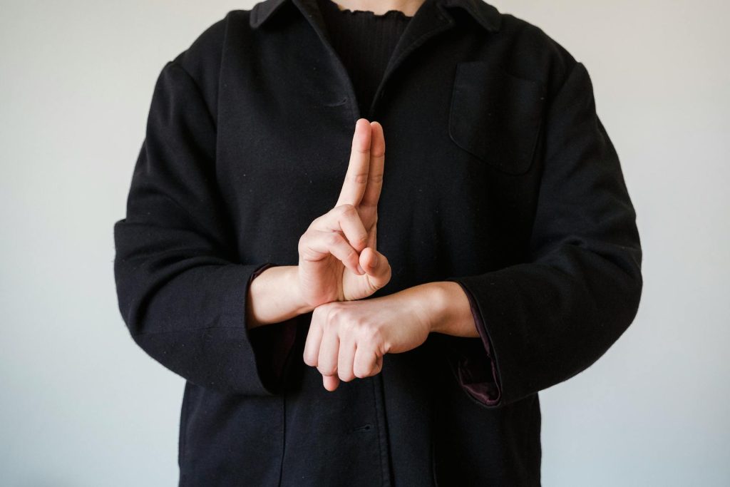 Eine Person zeigt eine Handgeste mit einem Finger, der aufrecht steht, und einer anderen Hand, die in einer anderen Position ist. Die Person trägt einen schwarzen Mantel und steht vor einem neutralen Hintergrund.