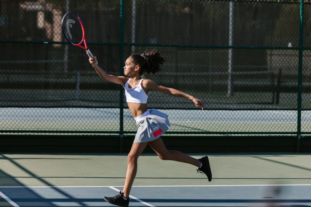 Ein Mädchen spielt Tennis auf einem Platz, während sie mit einem Schläger einen Ball schlägt. Sie trägt ein sportliches Outfit und zeigt eine dynamische Bewegung. Im Hintergrund sind Tennisnetze und ein gepflegter Platz sichtbar.