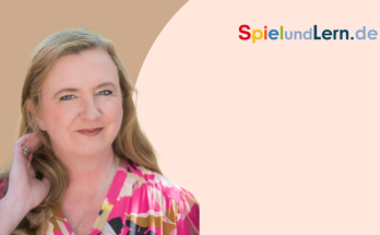 Porträt einer lächelnden Frau mit langen, blonden Haaren, die ein farbenfrohes, gemustertes Oberteil trägt. Im Hintergrund ist das Logo von SpielundLern.de zu sehen. Ideal für Inhalte über Bildung und spielerisches Lernen.