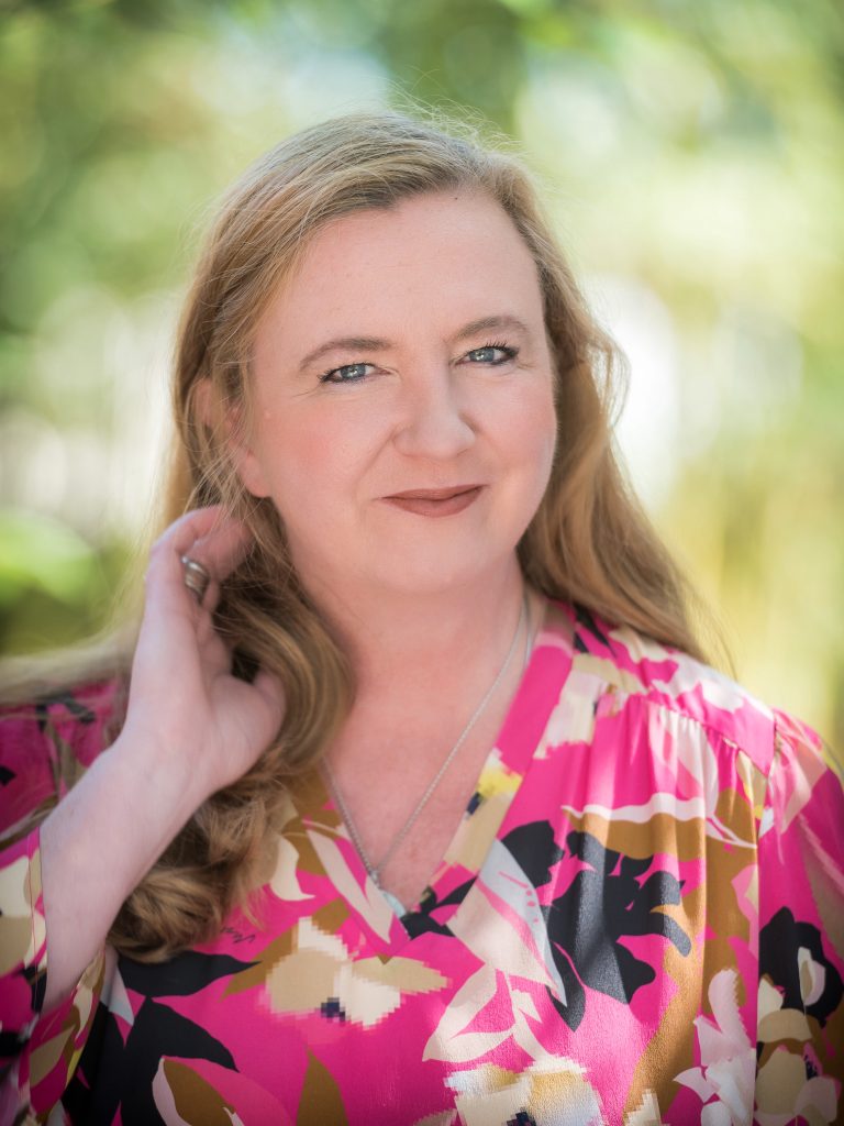 Porträt einer lächelnden Frau mit langen, lockigen, blonden Haaren, die ein farbenfrohes, blumenmusterndes Oberteil trägt. Der Hintergrund ist unscharf und zeigt eine natürliche, grüne Umgebung.