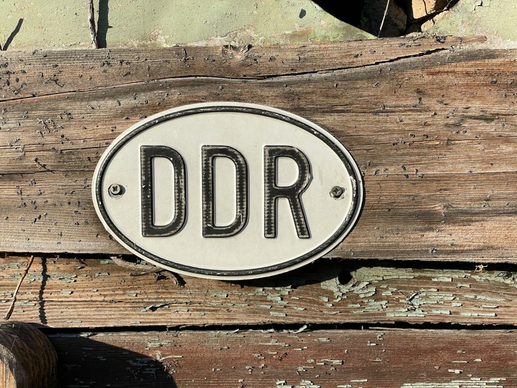 Schild mit der Aufschrift "DDR" auf verwittertem Holz, das die historische Bedeutung der Deutschen Demokratischen Republik symbolisiert.
