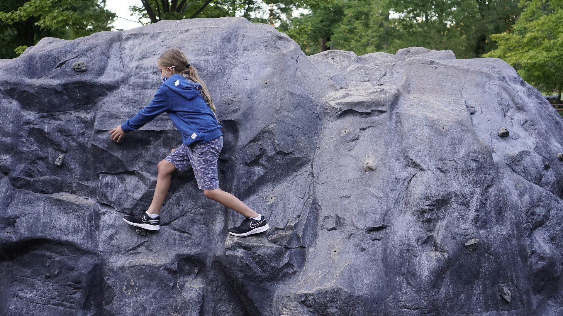 Ein Kind klettert an einem künstlichen Felsen im Freien. Es trägt einen blauen Hoodie und kurze Hosen, während es konzentriert nach Halt sucht. Die Umgebung ist grün und lädt zum Spielen ein.