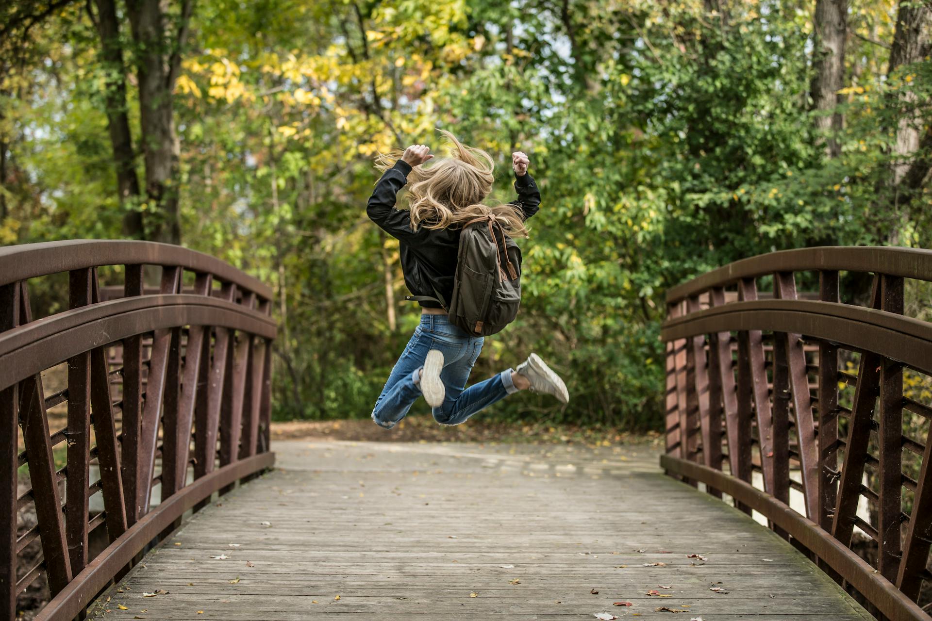 Ein Mädchen springt fröhlich auf einer Holzbrücke in einem herbstlichen Wald. Sie trägt eine schwarze Jacke und einen Rucksack, während bunte Blätter im Hintergrund zu sehen sind.