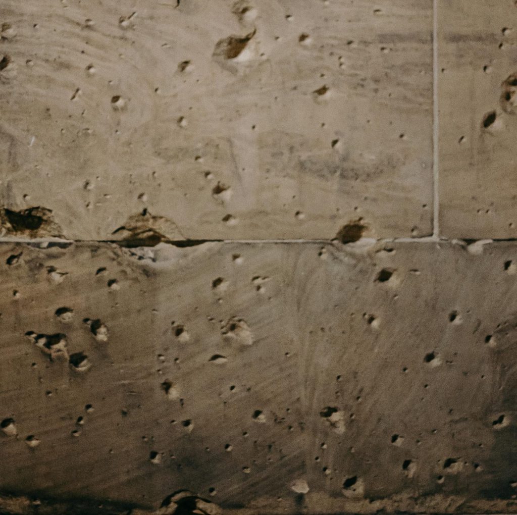 Texturierte Wand mit sichtbaren Einschlägen und Unebenheiten, die auf Alter und Abnutzung hinweisen. Die Oberfläche zeigt eine Mischung aus glatten und rauen Bereichen. Ideal für historische oder architektonische Themen.