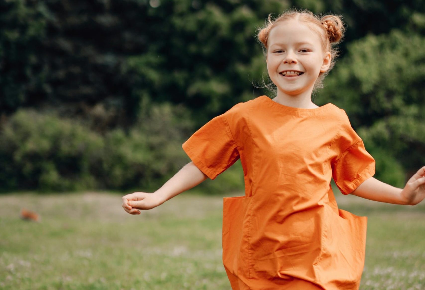 Ein fröhliches Mädchen in einem orangefarbenen Kleid läuft lachend über eine grüne Wiese, umgeben von Bäumen und einem blauen Himmel. Ideal für Themen wie Kinderbekleidung, Outdoor-Aktivitäten und Freude am Spiel.