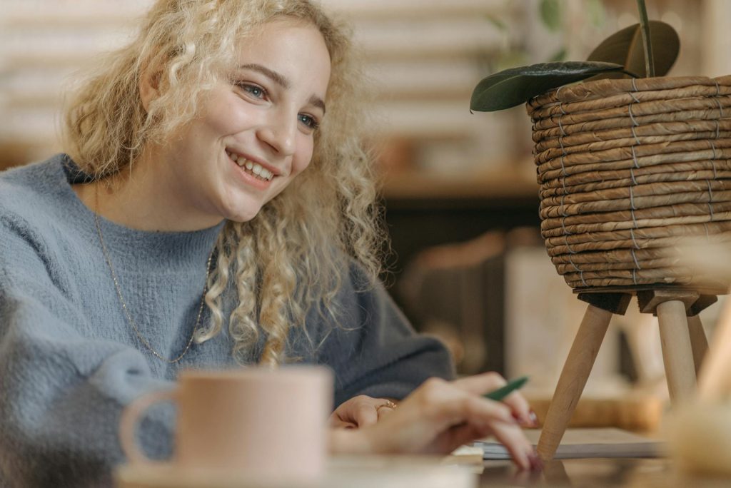 Junge Frau mit lockigem, blondem Haar lächelt, während sie an einem Tisch sitzt und Notizen macht. Im Hintergrund ist ein dekorativer Blumentopf sichtbar. Die Szene vermittelt eine kreative und entspannte Atmosphäre.